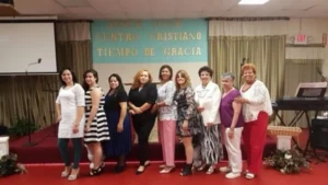 A group of nine women standing in front of a church stage with a sign reading "Jesús Vive Centro Cristiano Tiempo de Gracia