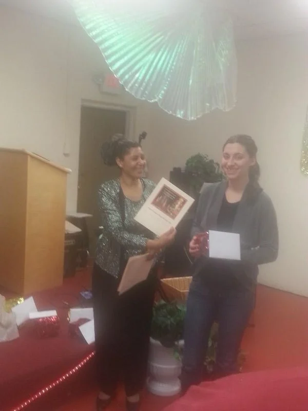 Two women stand indoors, one holding a certificate and the other a small box