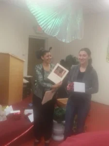 Two women stand indoors, one holding a certificate and the other a small box