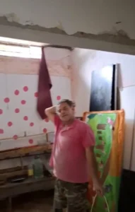 A man stands in a room with a pink wall, dressed casually, with a neutral expression on his face.