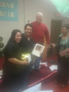 Three individuals stand in front of a piano, each holding a wrapped gift, smiling at the camera.