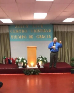 A man speaks on a stage with a podium and plants. Behind him is a sign reading "Jesús Vive Centro Cristiano Tiempo de Gracia.